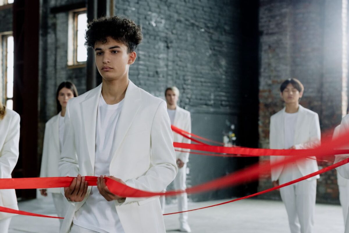 Group of young fashion models in white suits with a red ribbon in an industrial setting.