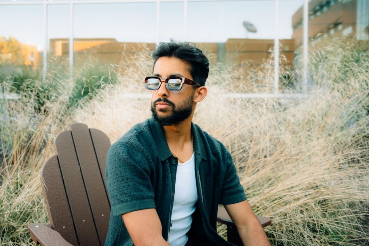 Fashionable man with beard sitting outdoors on a wooden chair, wearing sunglasses and a green shirt.